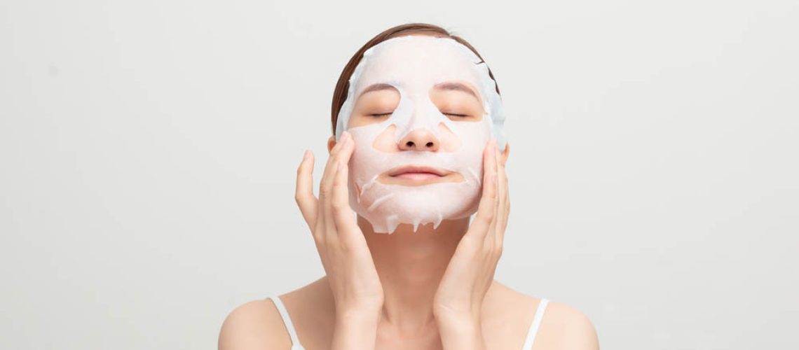 Beautiful asian woman applying paper sheet mask on her face white background